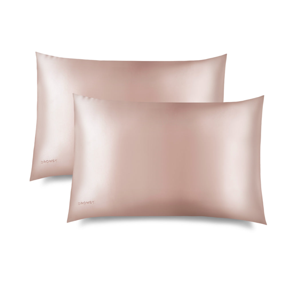 Two Sunset Pink Silk Pillowcases by us.drowsysleepco, featuring a smooth, shiny finish, are shown overlapping on a white background—an ideal addition to your skincare routine and perfect for protecting hair as you sleep.