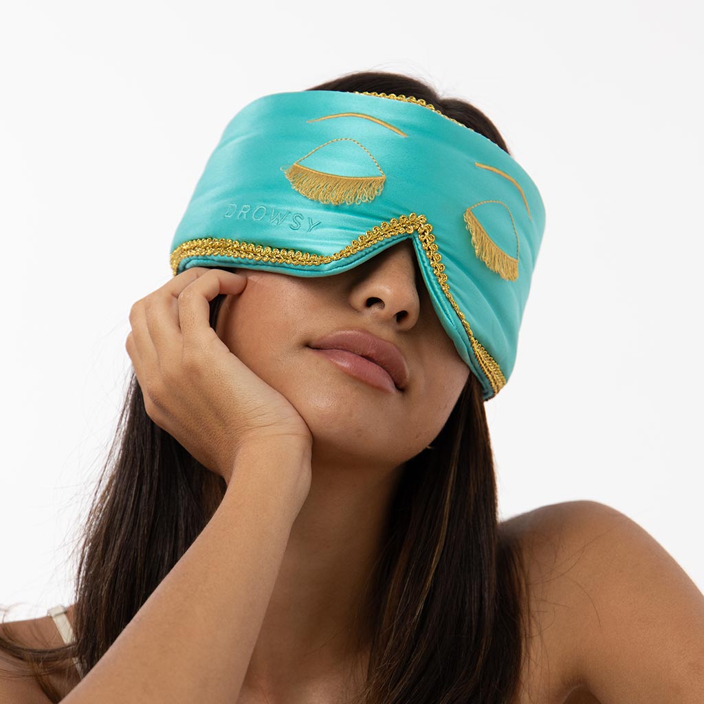 A woman with long brown hair looks relaxed against a white background, wearing the drowsysleepco Breakfast at Drowsy Eyelash Protecting Mask—a teal sleep mask with gold trim and embroidered closed eyes.