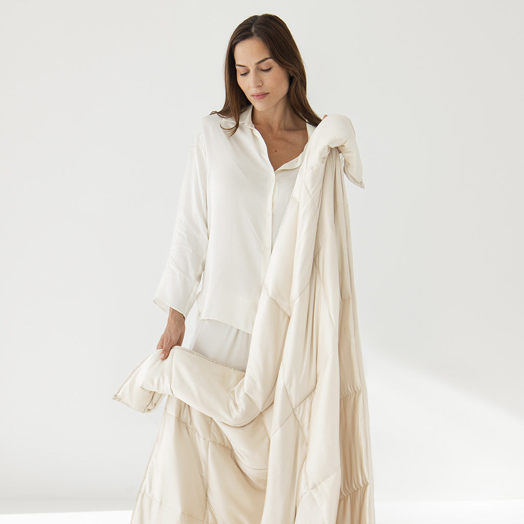 A woman in a loose white shirt gently drapes the us.drowsysleepco Dusty Gold Silk Weighted Blanket, its cream-colored silk cover offering a sense of deep relaxation against a plain white background.