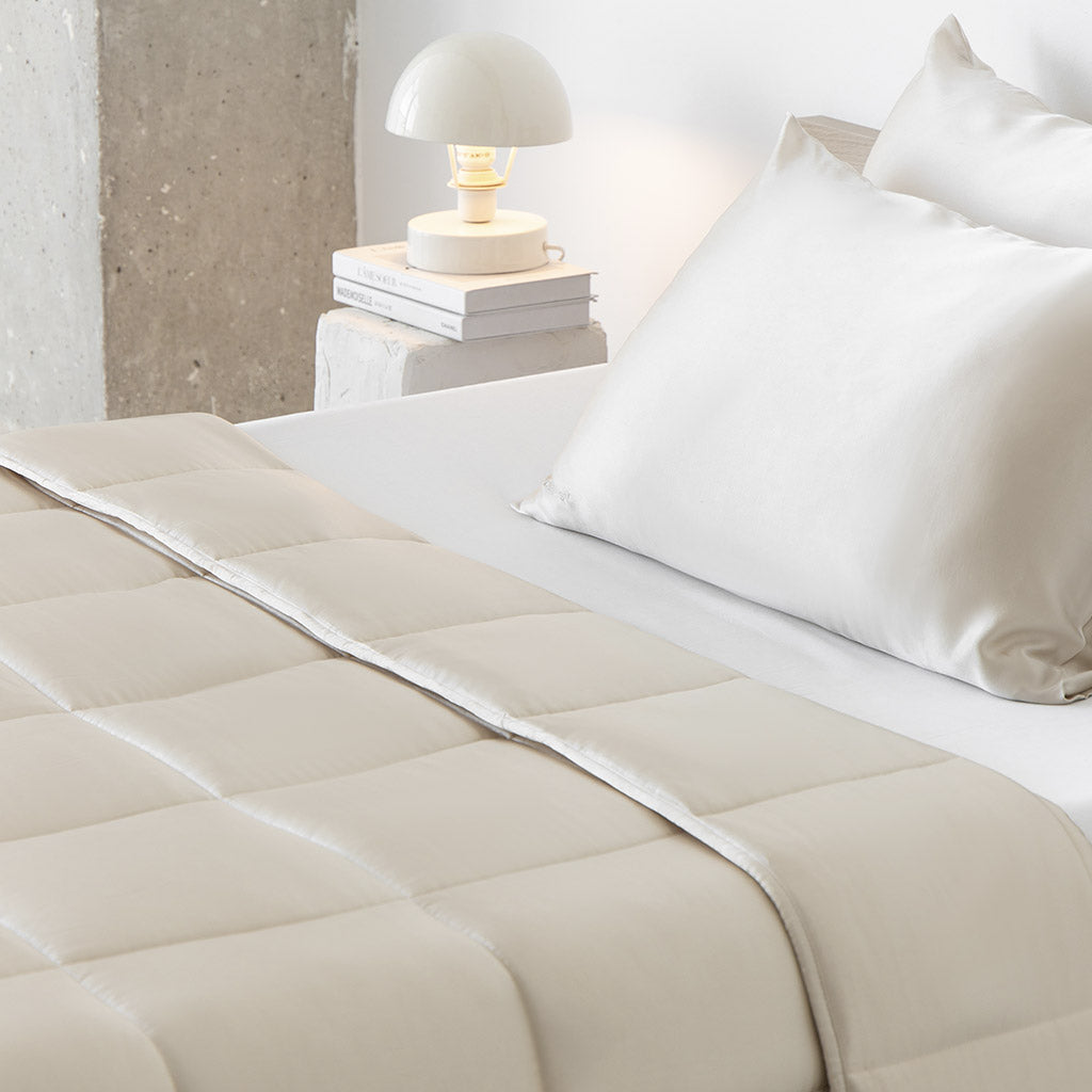 A neatly made bed with white sheets and pillows is topped with the us.drowsysleepco Dusty Gold Silk Weighted Blanket. A small lamp and stacked books on the bedside table complete this calm, modern space for deep relaxation.