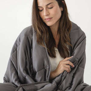 A woman with long brown hair, wearing a white top, relaxes with her eyes closed while wrapped in the Moonlight Shadow Silk Weighted Blanket by us.drowsysleepco, against a plain light background.