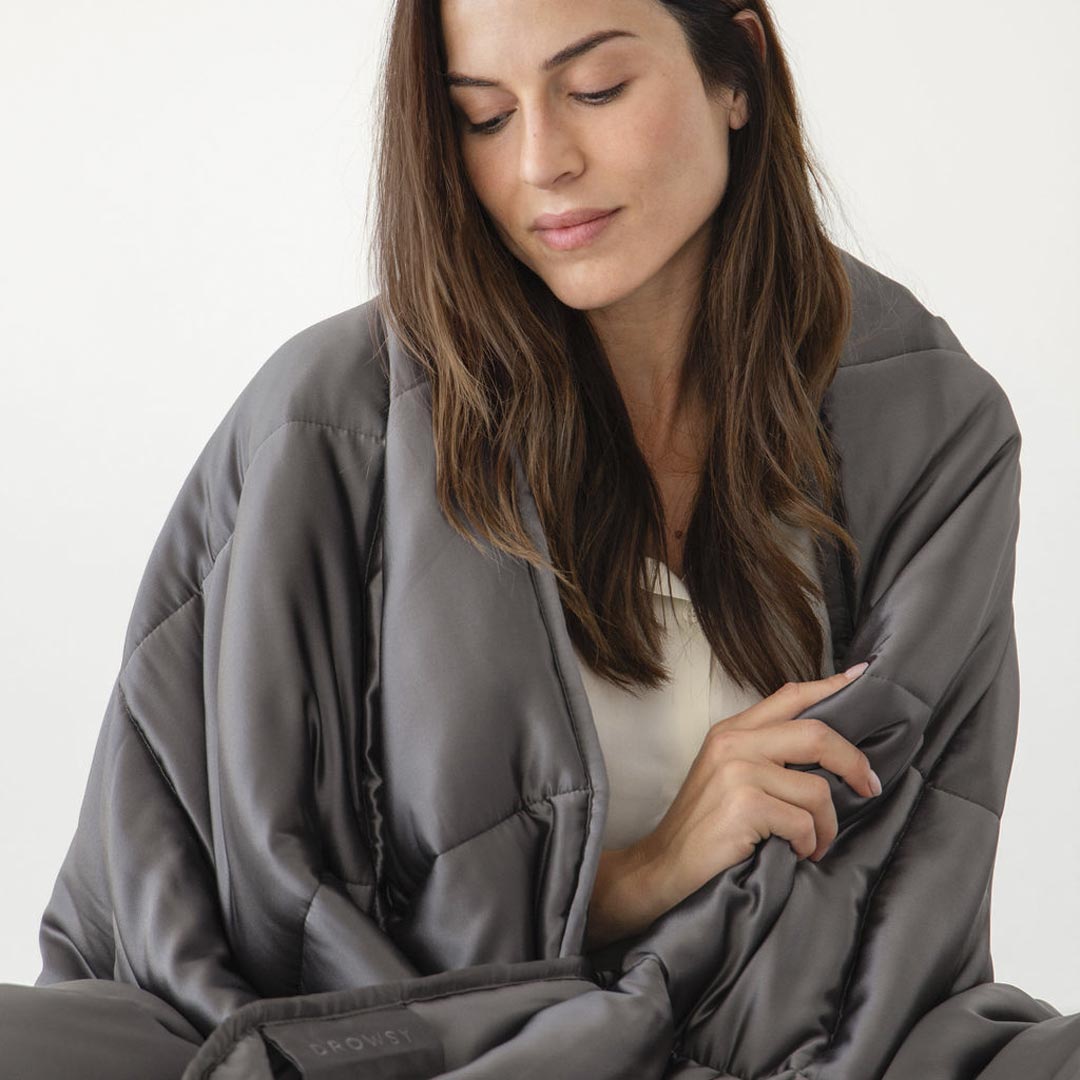 A woman with long brown hair, wearing a white top, relaxes with her eyes closed while wrapped in the Moonlight Shadow Silk Weighted Blanket by us.drowsysleepco, against a plain light background.