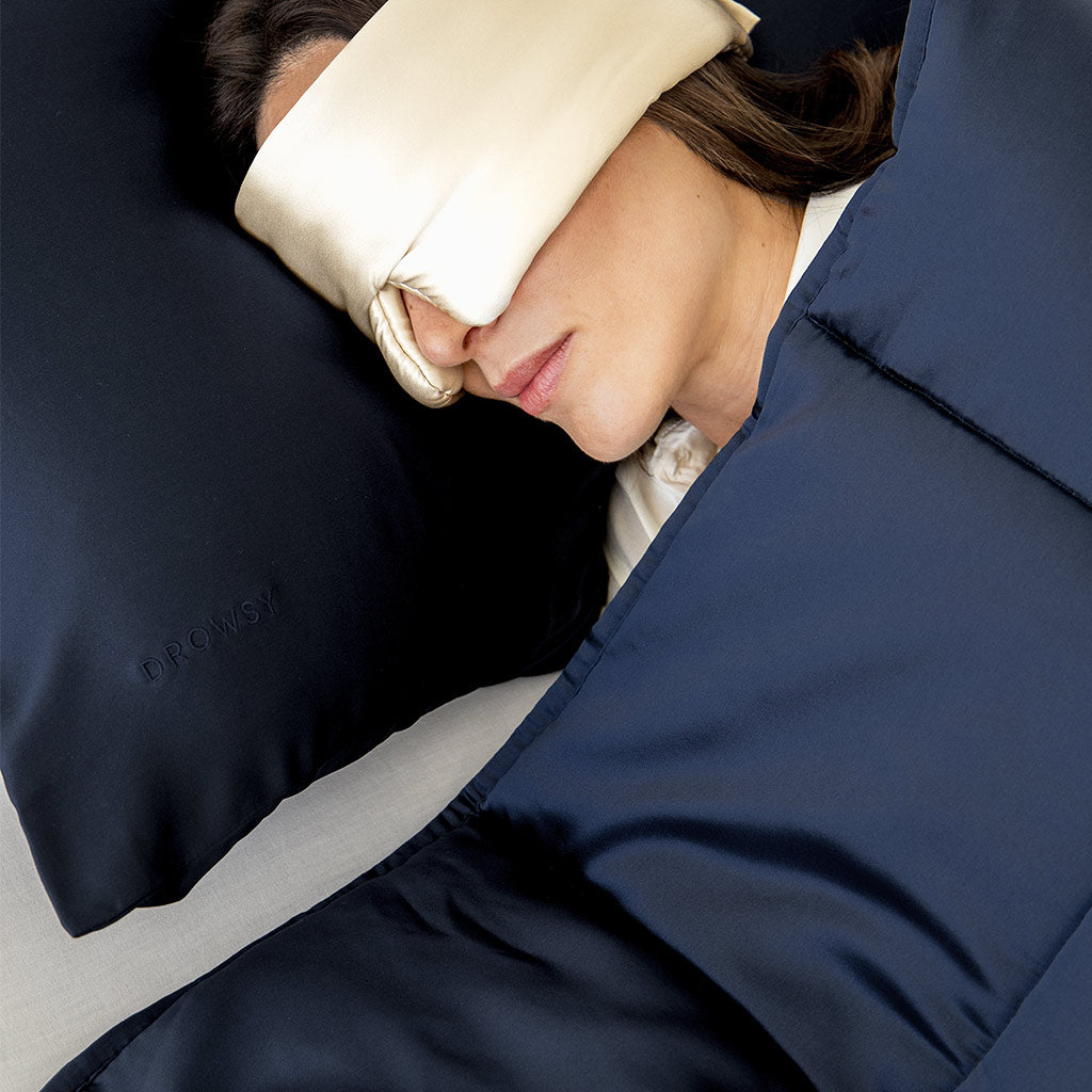 A person rests with a cream silk sleep mask, lying on a navy pillow embroidered with DROWSY and covered by the us.drowsysleepco Midnight Blue Silk Weighted Blanket, creating an atmosphere of deep relaxation.