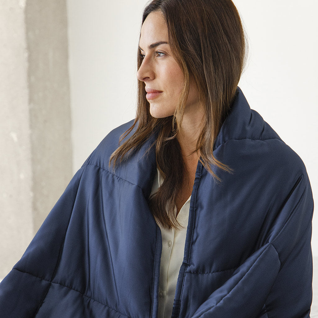 A woman with long brown hair, wrapped in the us.drowsysleepco Midnight Blue Silk Weighted Blanket and wearing a light top, gazes to the side in softly lit indoor surroundings.
