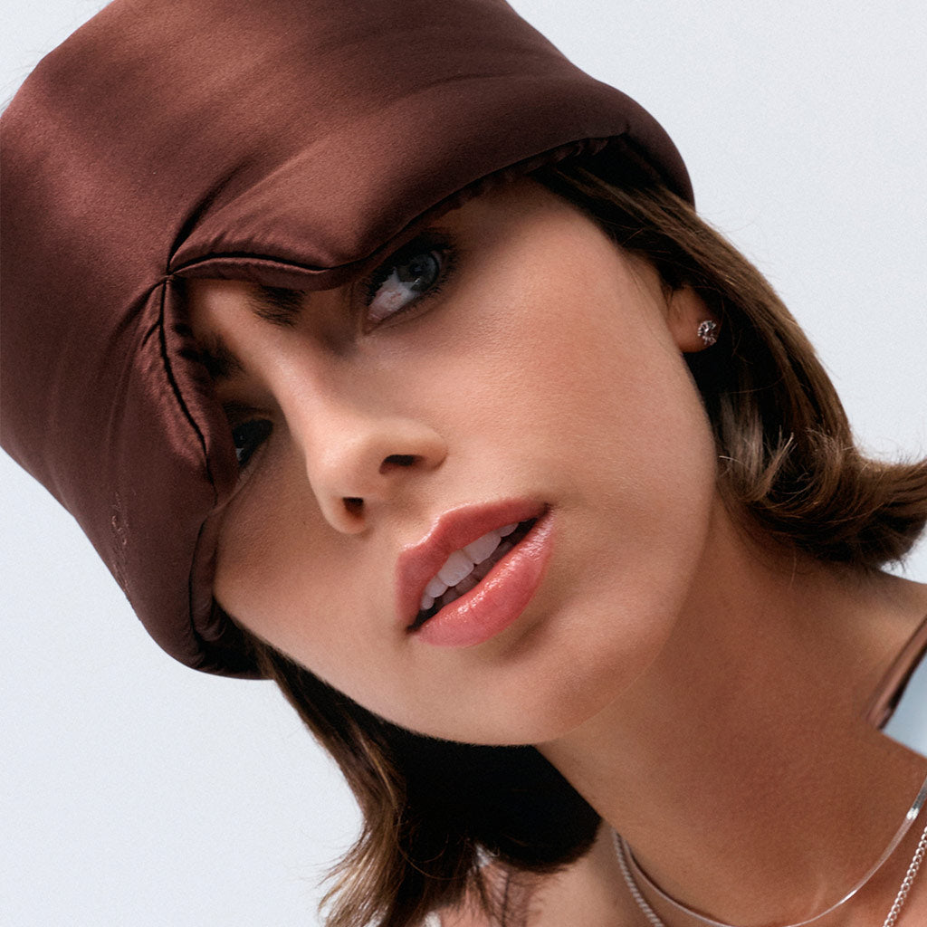 A woman with short brown hair wears the drowsysleepco Cocoa Glow sleep mask, styled chicly over one eye like a shiny brown hat. She sports light makeup, pink lipstick, small earrings, and a silver necklace against a light background.