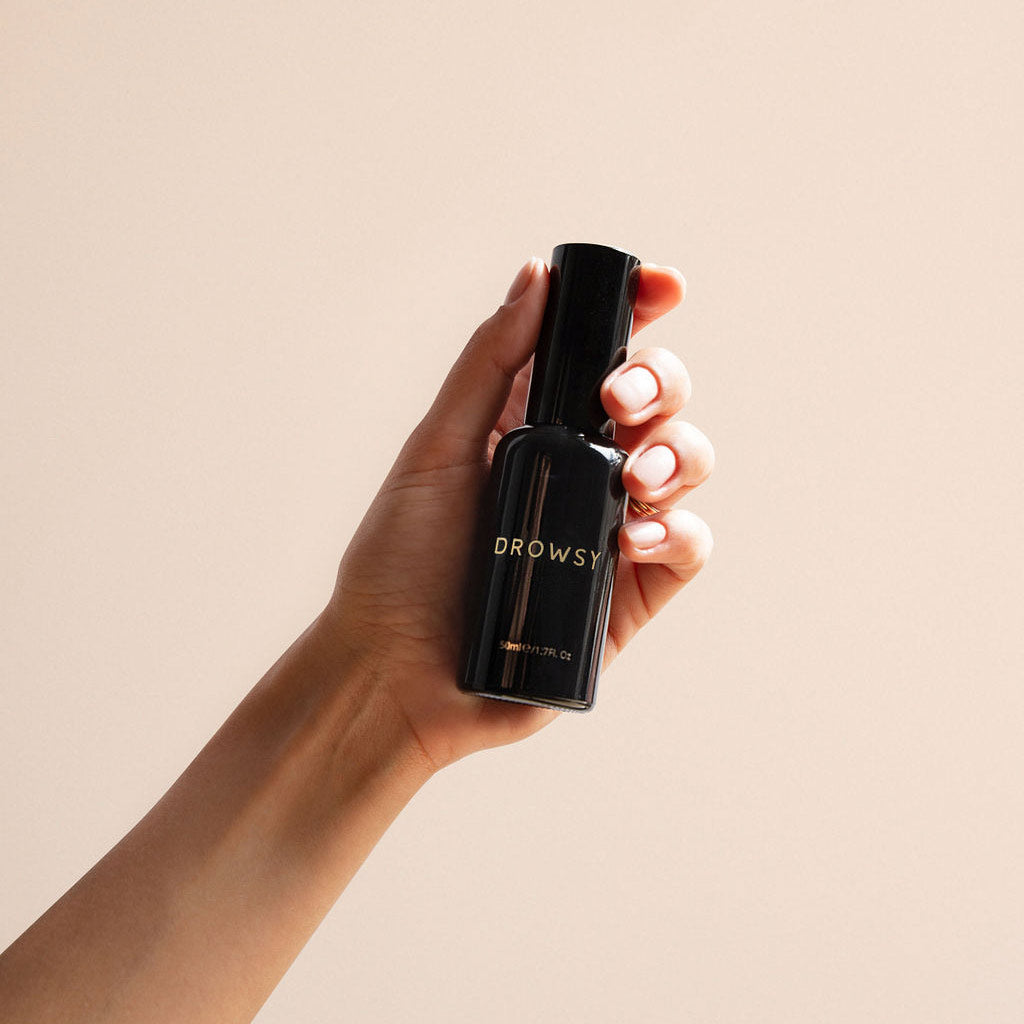 A hand holds a sleek black bottle labeled DROWSY against a neutral beige background.