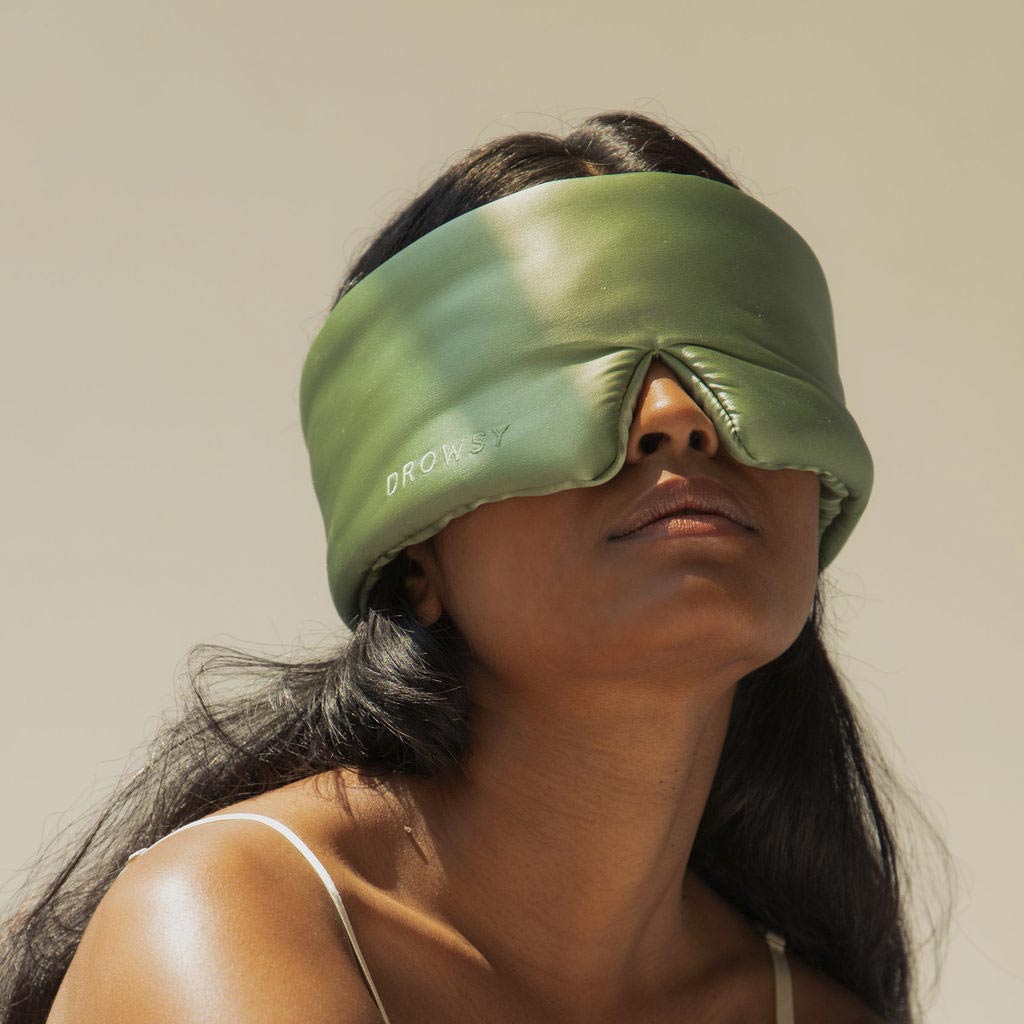A woman with long dark hair wears a large, green silk sleep mask labeled DROWSY. She is looking slightly upward, with a relaxed expression against a plain, light background.