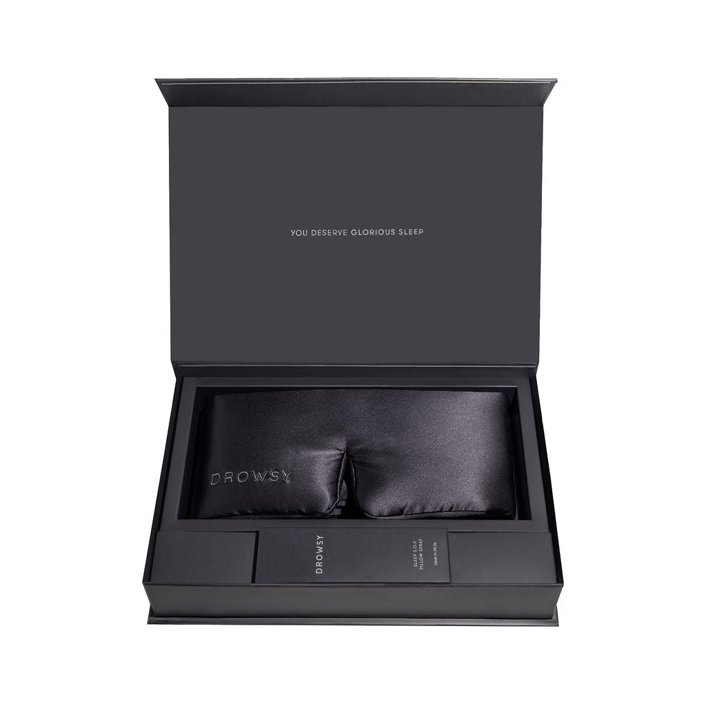 A black gift box with You deserve glorious sleep inside the lid, containing a black silk sleep mask and a slim black case, both labeled Drowsy.
