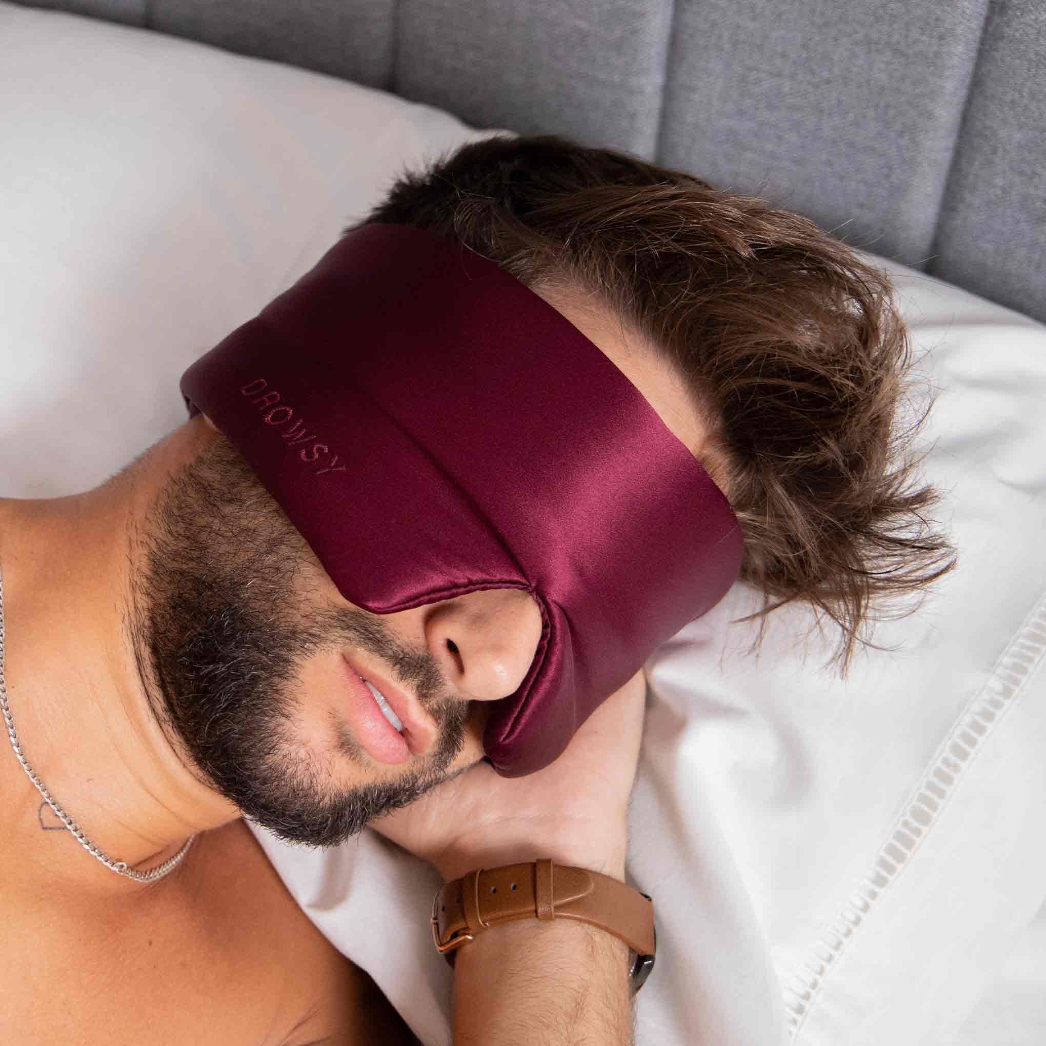 A man with a beard is lying on his side in bed, wearing a burgundy silk sleep mask. The background shows a white pillow and a grey upholstered headboard.
