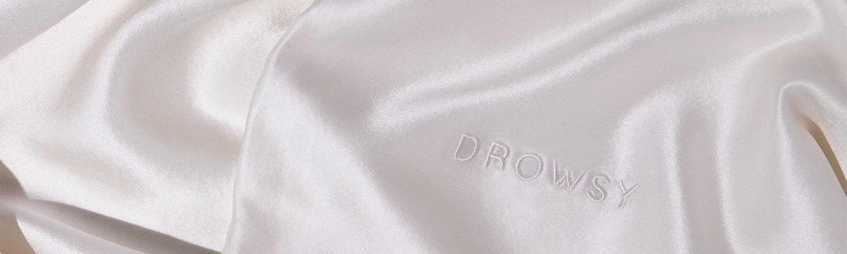 Close-up of smooth, shiny, light-colored satin fabric with the word DROWSY embroidered in matching thread on the surface.