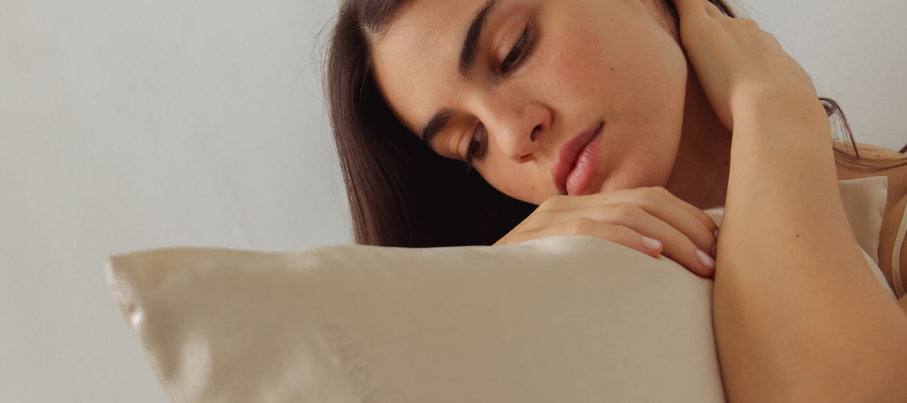 A young woman with long dark hair gently rests her head and hand on a light-colored satin pillow, looking downwards with a calm, thoughtful expression.