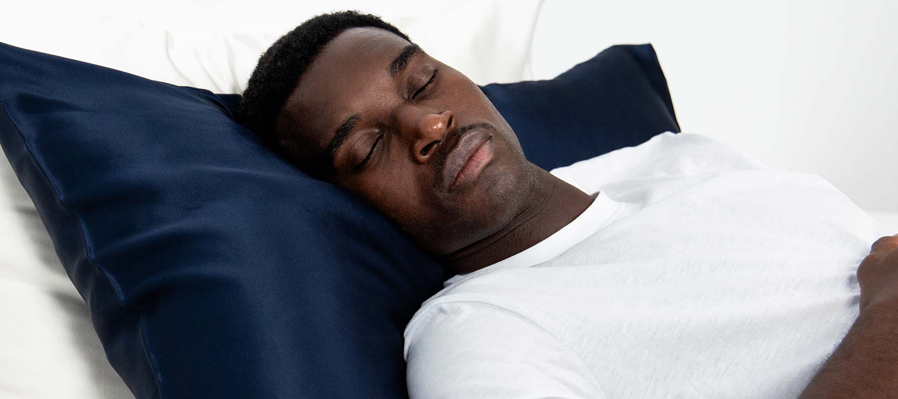 A man lies on his back in bed, eyes closed and appearing asleep, resting his head on the drowsysleepco Midnight ritual bundle in Midnight Blue silk pillowcase.