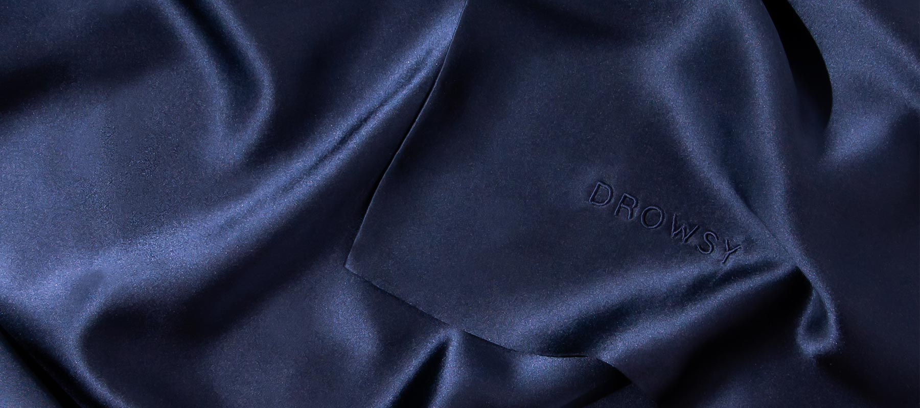 A close-up of smooth, dark navy blue silk fabric with the word “DROWSY” subtly embossed into the material. The fabric has a soft sheen and gentle folds.