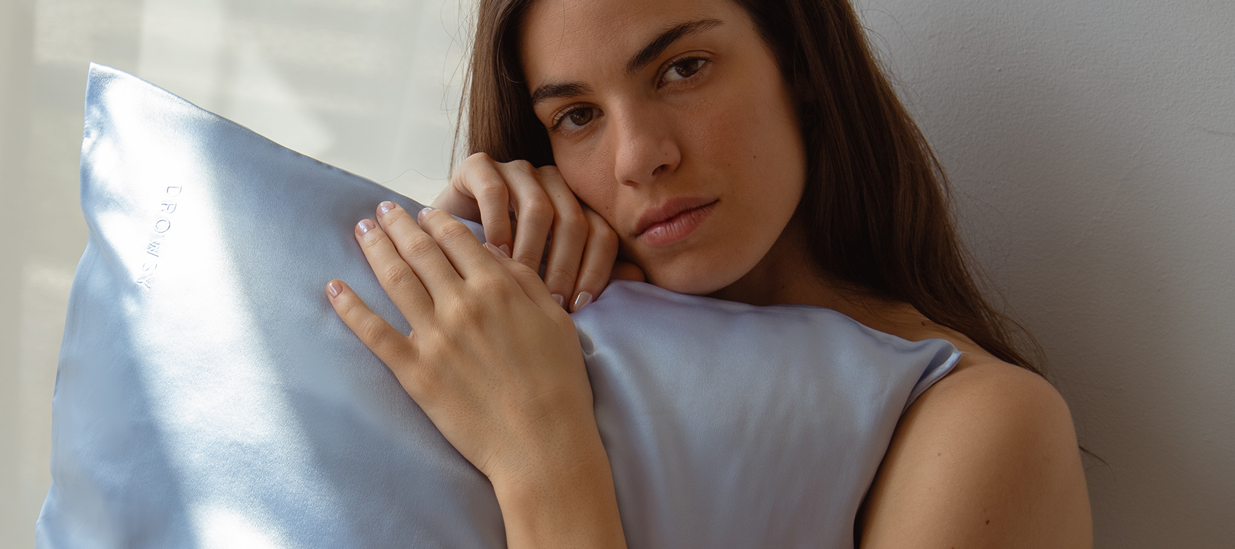 A woman with long dark hair hugs a shiny light blue pillow, resting her face and hands on it, while looking softly at the camera in natural light.