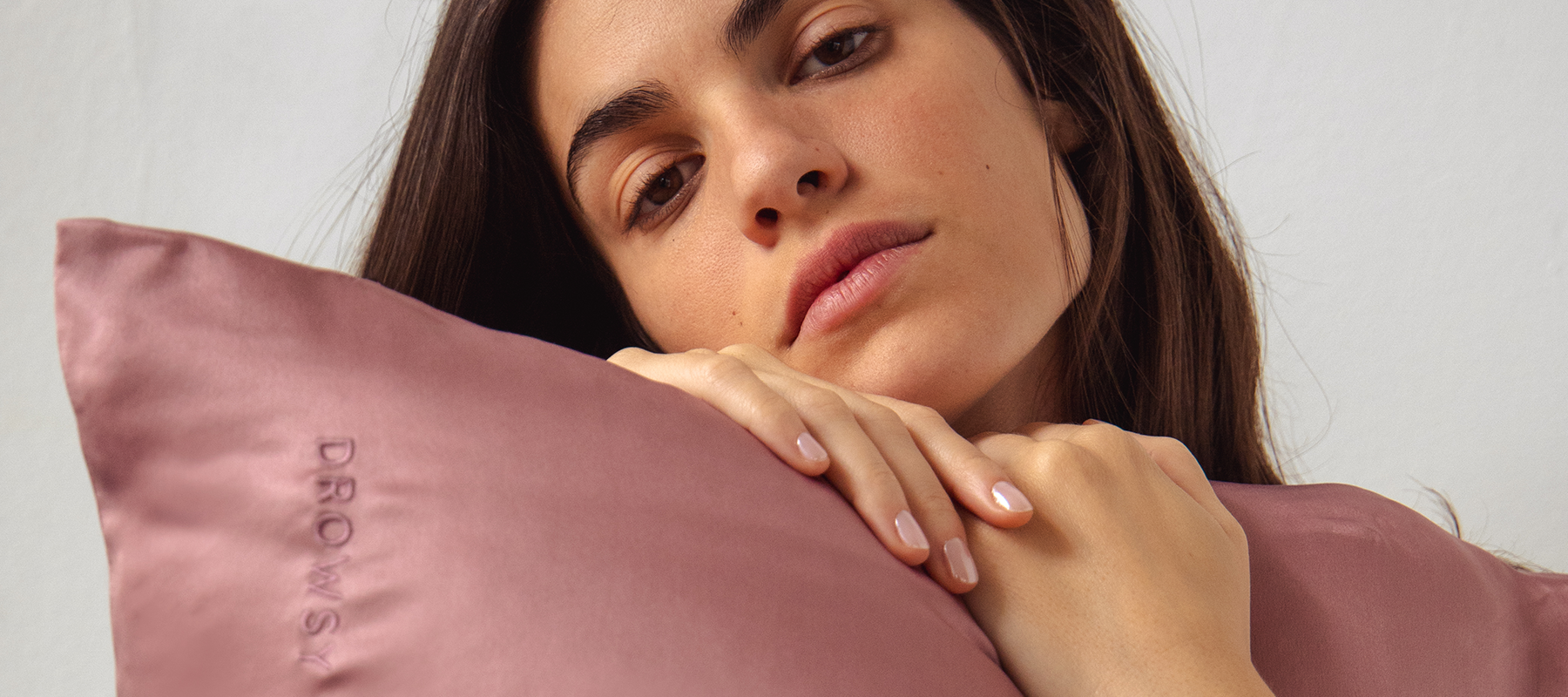 A woman with long brown hair rests her chin and hands on a mauve satin pillow embroidered with the word DROWSY, gazing softly at the camera against a plain light background.