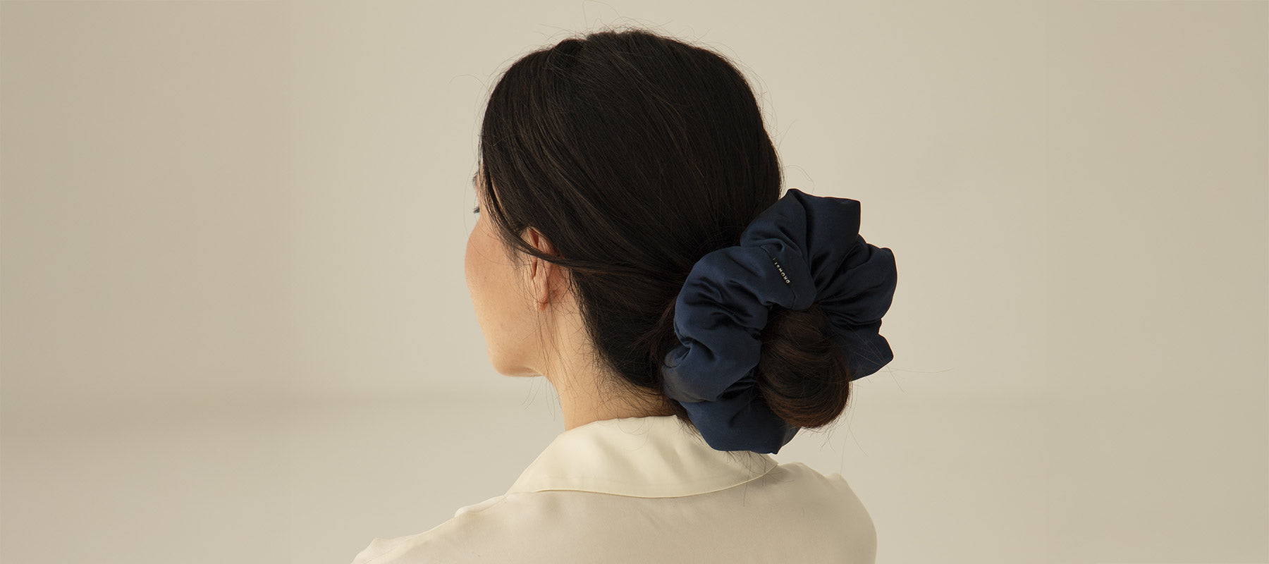 A person with dark hair styled in a low bun wears a large navy blue scrunchie and a cream-colored top, facing away from the camera against a plain, light background.
