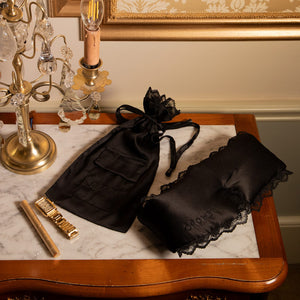 A marble-top table displays the drowsysleepco Paris Love Story - Black Jade Sleep Mask + Pouch, with lace trim and anti-aging benefits, beside a gold candle holder, gold comb, and an ornate lamp in warm light.