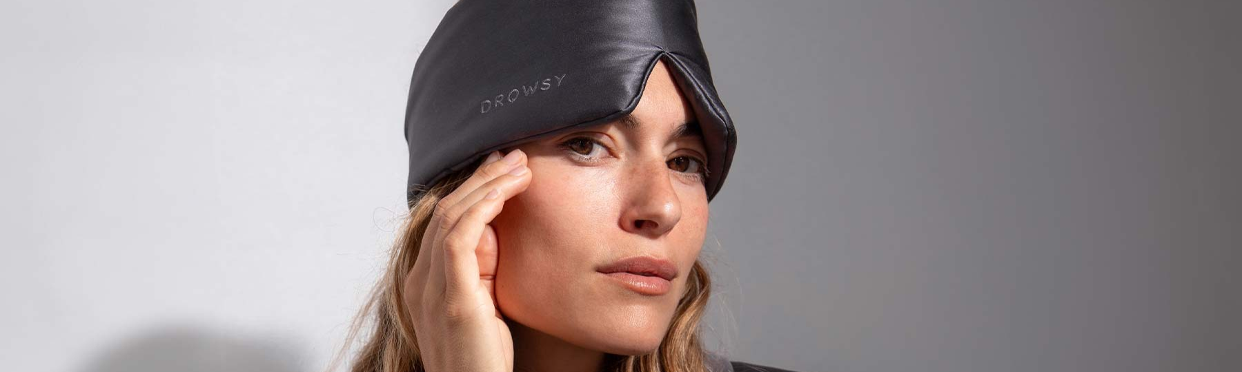 A woman with long hair wears a black sleep mask labeled DROWSY on her forehead, with one hand raised to her temple, against a plain background.