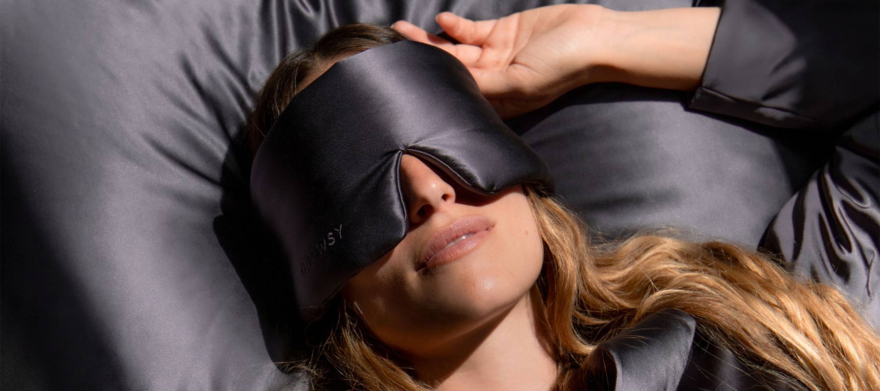 A person with long hair is lying on a bed, wearing a black silk eye mask and matching sleepwear, resting on a silky pillowcase in a softly lit setting.