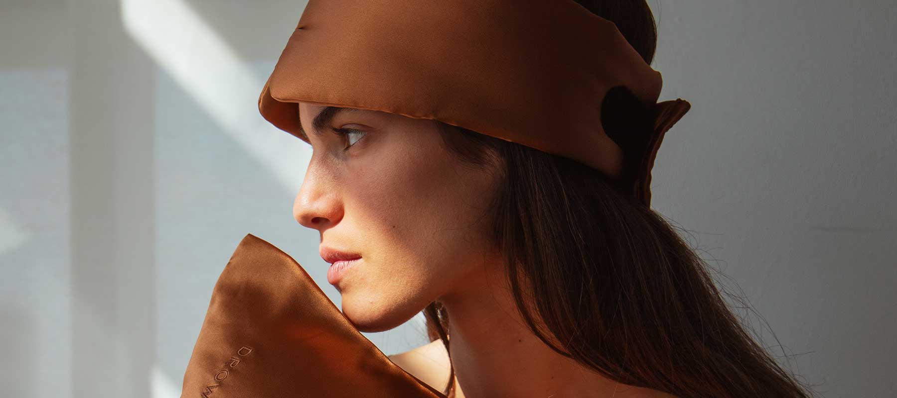 A person with long brown hair wears a brown silk eye mask pushed up on their forehead and holds a matching silk pillow, looking thoughtfully to the side in soft natural light.