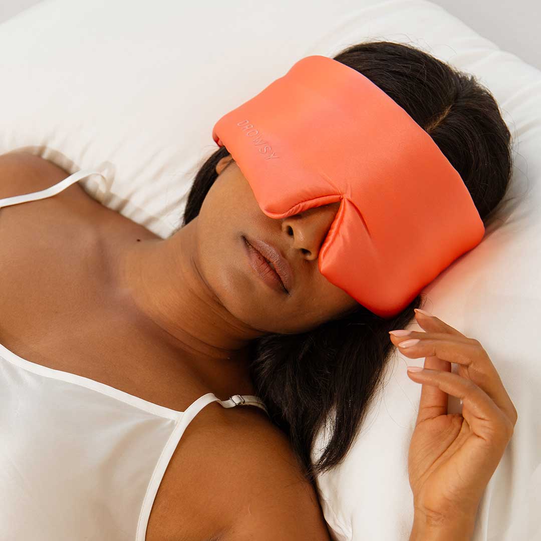 A woman lying on a white pillow wears an orange silk eye mask while resting, dressed in a white camisole.