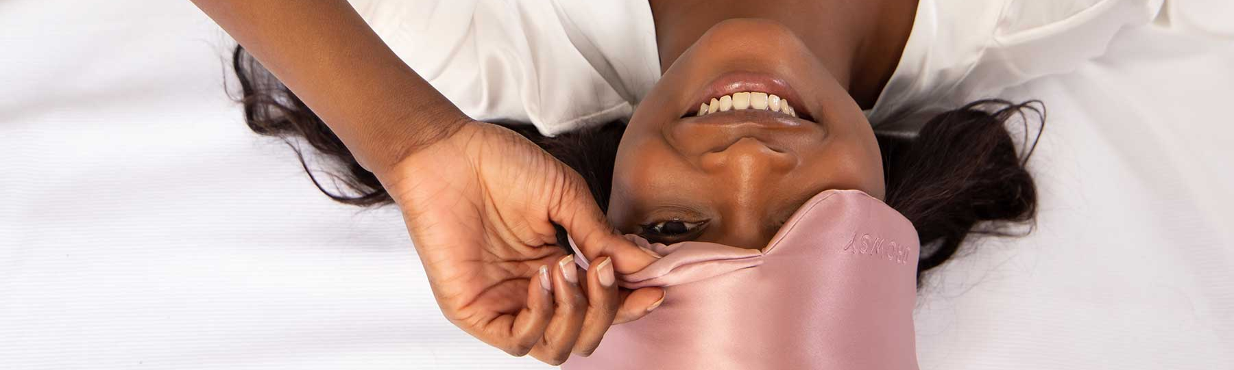 A woman lying on her back on a white bed, smiling and partially removing a pink satin sleep mask from her eyes.