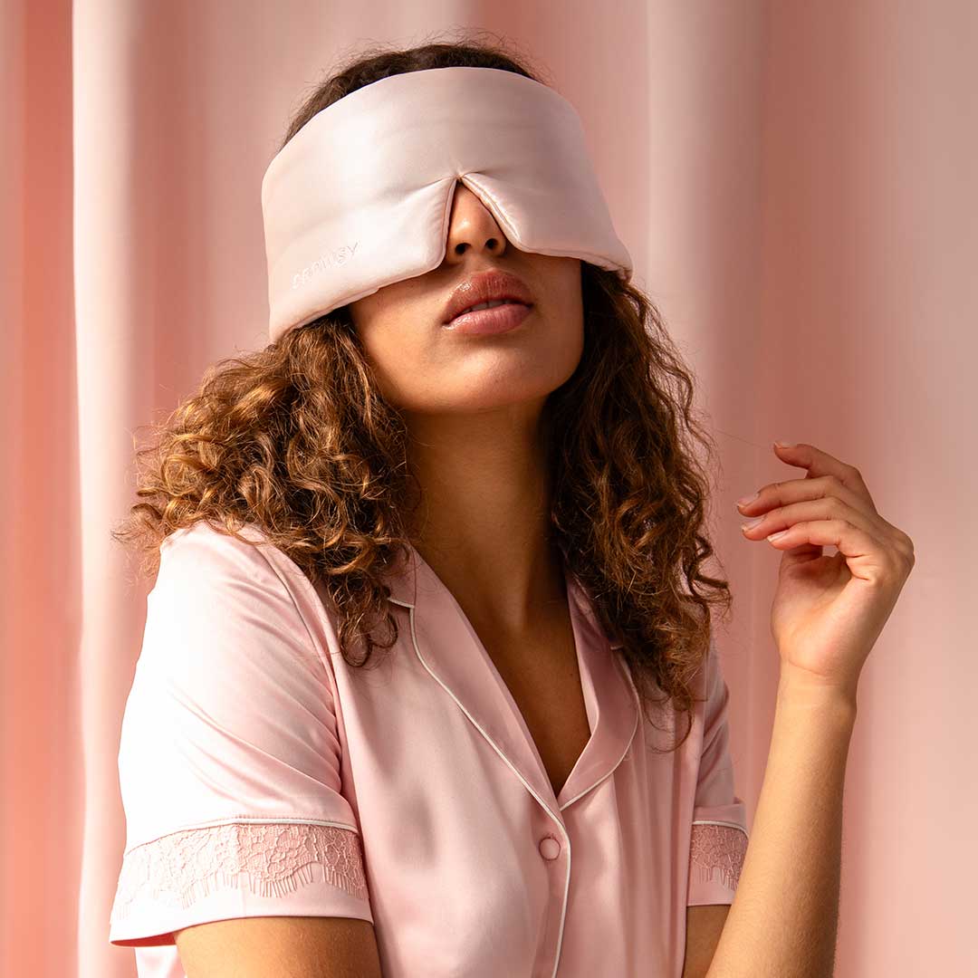 A woman with curly hair wearing a light pink pajama top and a large satin sleep mask sits against a pink background, with her head slightly tilted and one hand raised near her face.