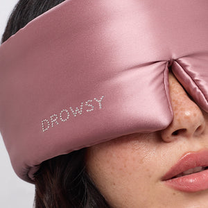 A close-up of a woman wearing the drowsysleepco Damask Rose sleep mask, featuring a Swarovski®-embellished logo, covering her eyes and part of her nose.