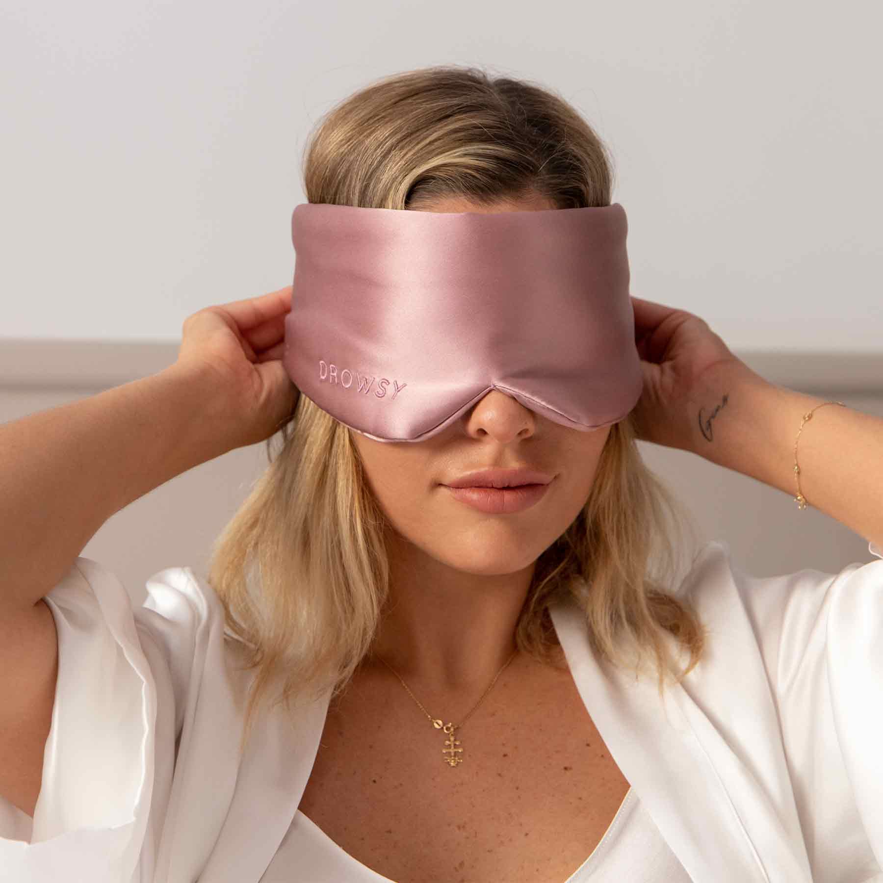 A woman with blonde hair in a white robe adjusts the Damask Rose silk eye mask by us.drowsysleepco, featuring DROWSY embroidery. The softly lit, neutral background enhances the eye mask’s relaxing appeal.