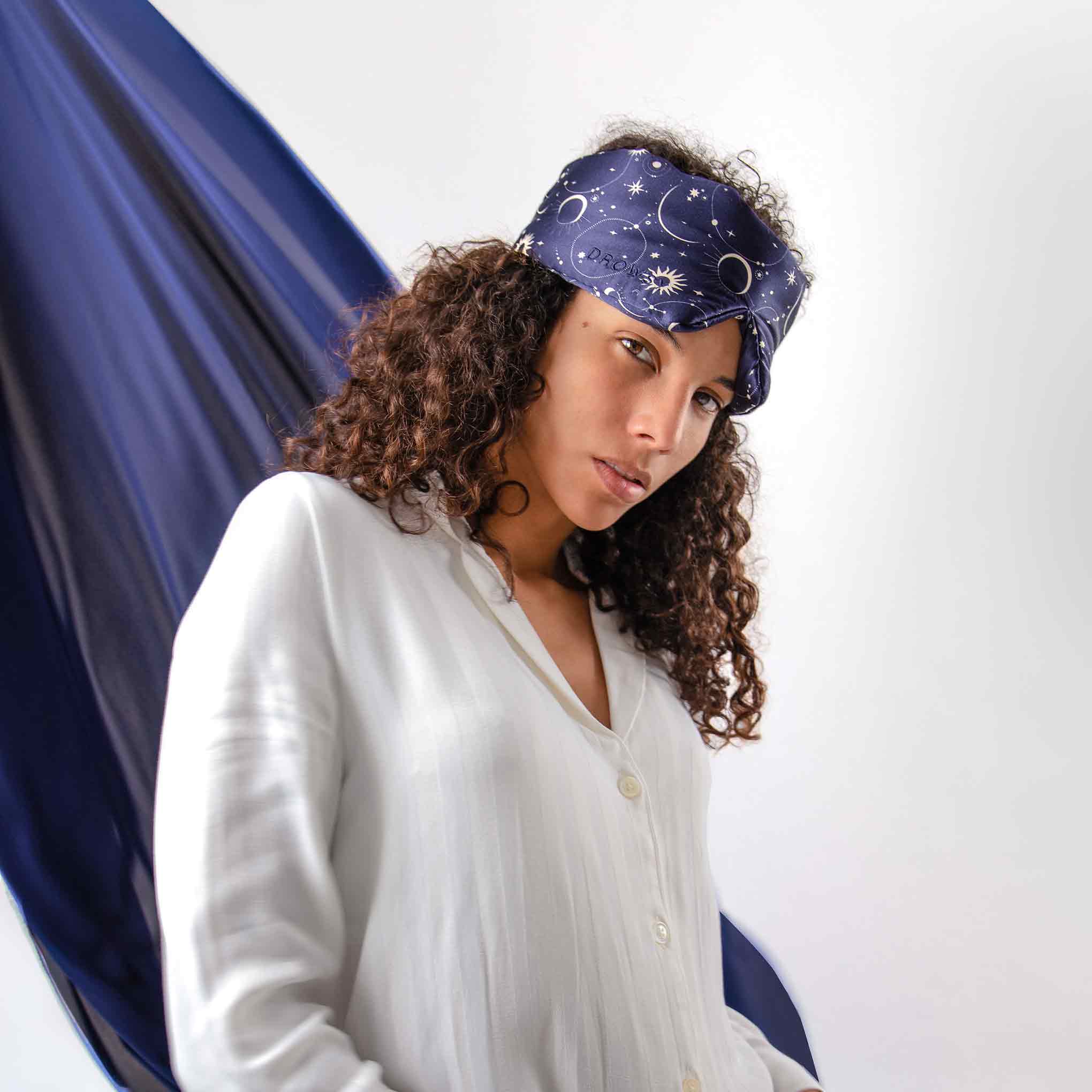 A woman with curly hair wears a white shirt and the “Cosmic Energy” navy silk eye mask by us.drowsysleepco, featuring celestial designs, as she stands before a plain background with flowing dark blue fabric behind her.