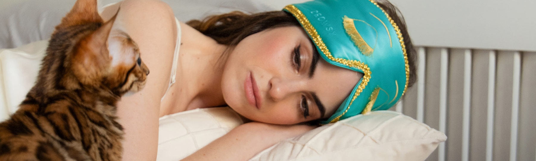 A woman wearing a teal and gold sleep mask rests on a pillow, eyes closed, while a brown striped cat sits nearby looking at her.