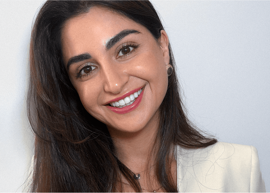 A woman with long dark hair smiles at the camera. She is wearing a light-colored blazer, a necklace, small earrings, and has natural makeup. The background is plain and light-colored.