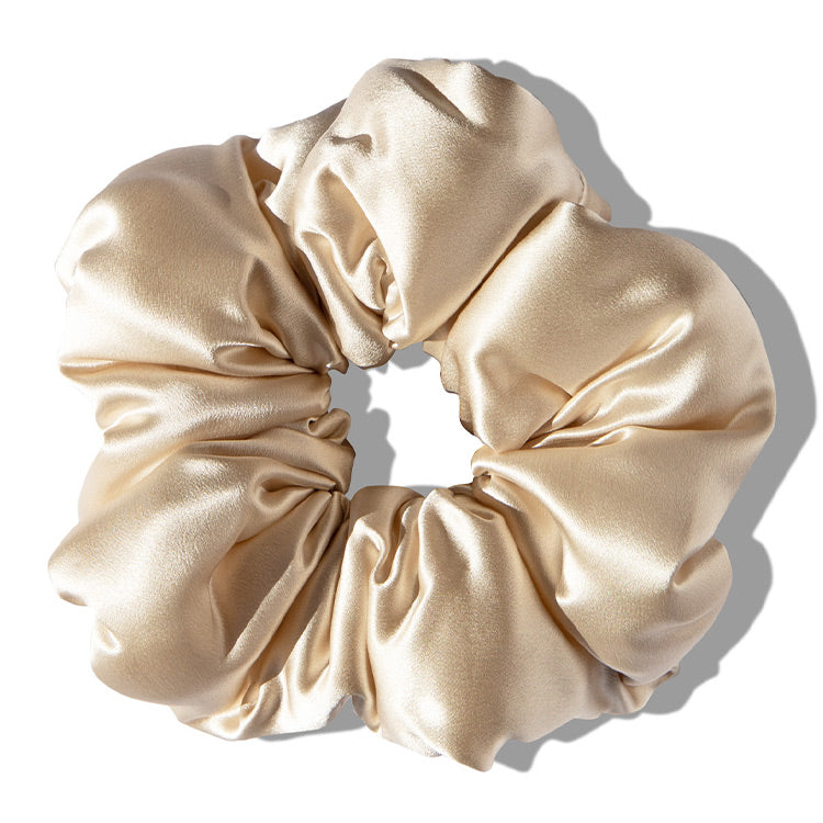 A shiny, light beige satin scrunchie with soft, gathered fabric arranged in a circular shape, casting a shadow on a white background.