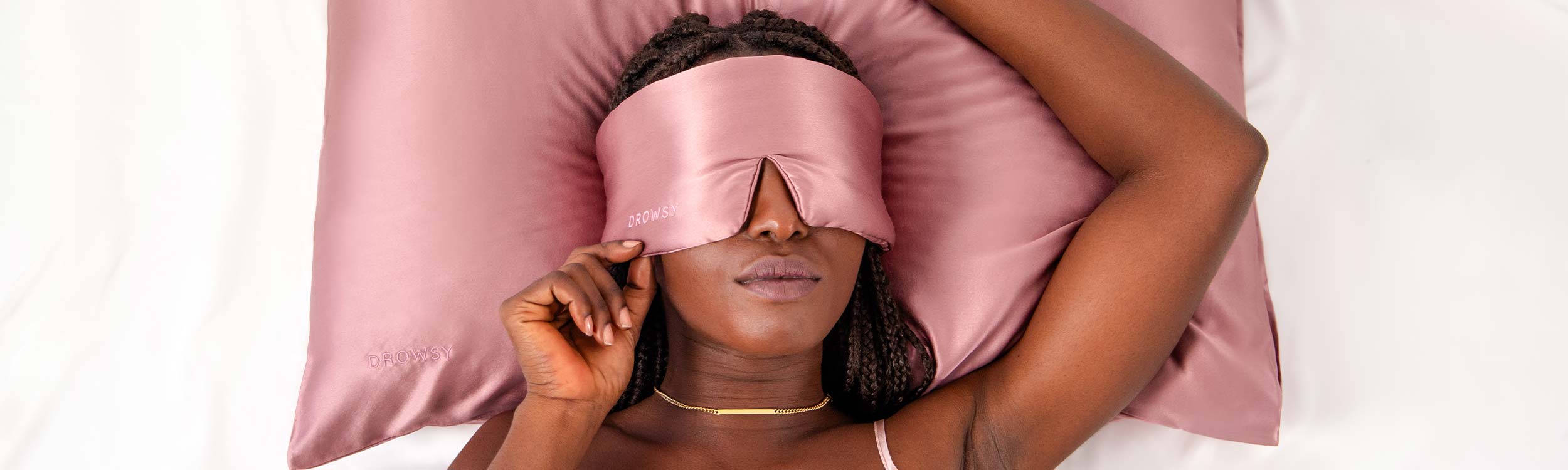 A woman with braided hair lies on a pink satin pillow, wearing a matching pink satin sleep mask and a gold necklace, resting her hand on the edge of the mask.