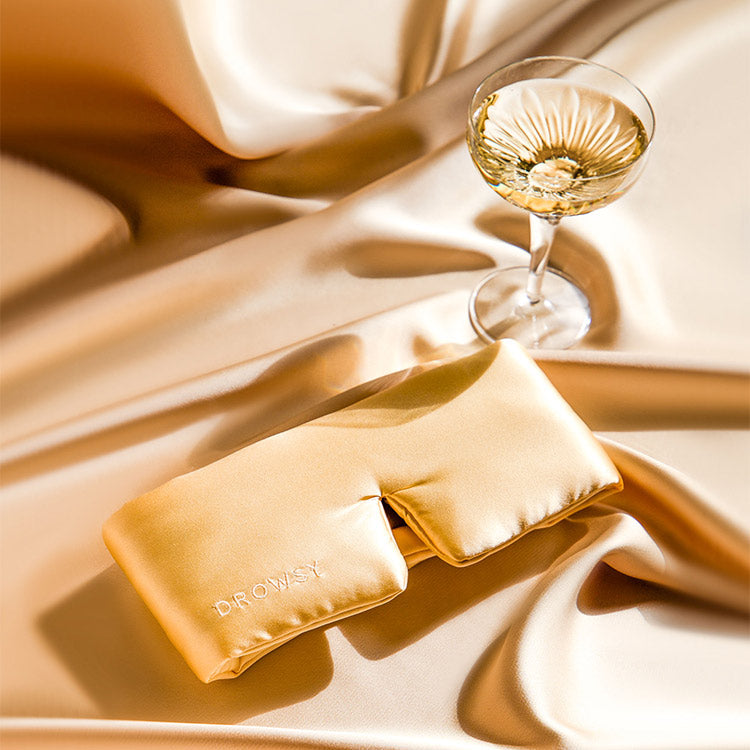 A golden satin eye mask embroidered with DR. OWISY lies on silky gold fabric beside a glass of white wine, creating a luxurious and relaxing atmosphere.