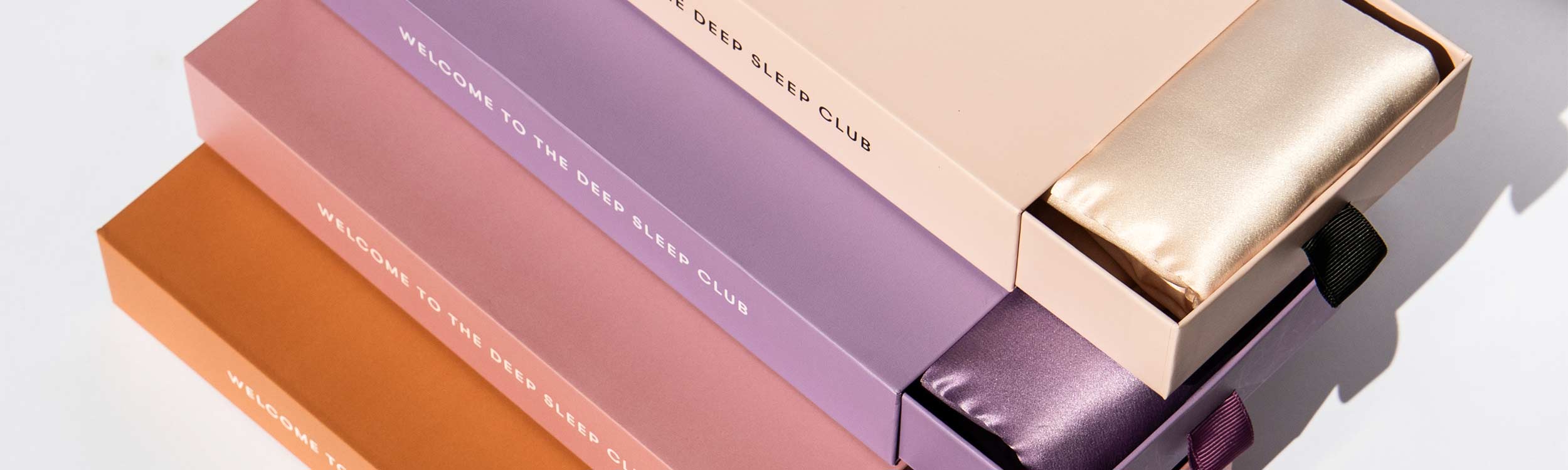 Four pastel-colored boxes, each labeled WELCOME TO THE DEEP SLEEP CLUB, are stacked diagonally. Two open boxes reveal shiny silk sleep masks—one gold, one purple—inside.