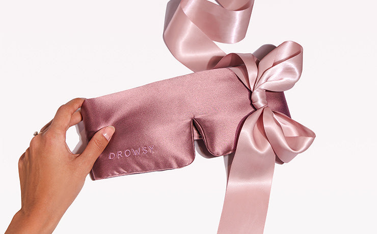 A hand holds a silky pink sleep mask with the word “DROWSY” embroidered on it, featuring a large matching satin ribbon tied in a bow.
