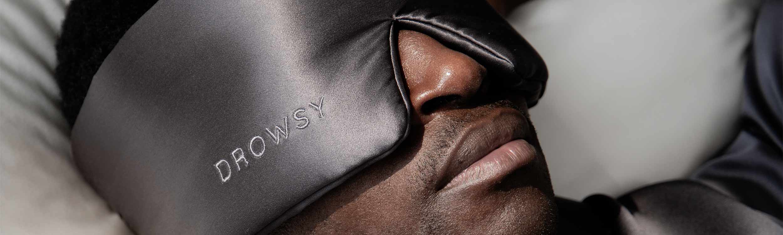 A close-up of a person wearing a dark satin sleep mask embroidered with the word DROWSY, lying on a pillow, suggesting restful sleep.