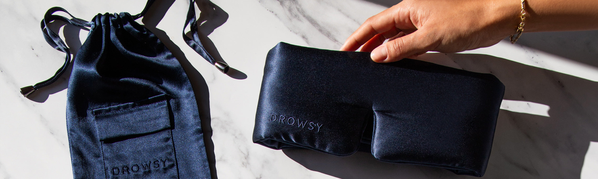 A hand holds a navy blue DROWSY sleep mask on a marble surface, with a matching fabric pouch lying nearby.