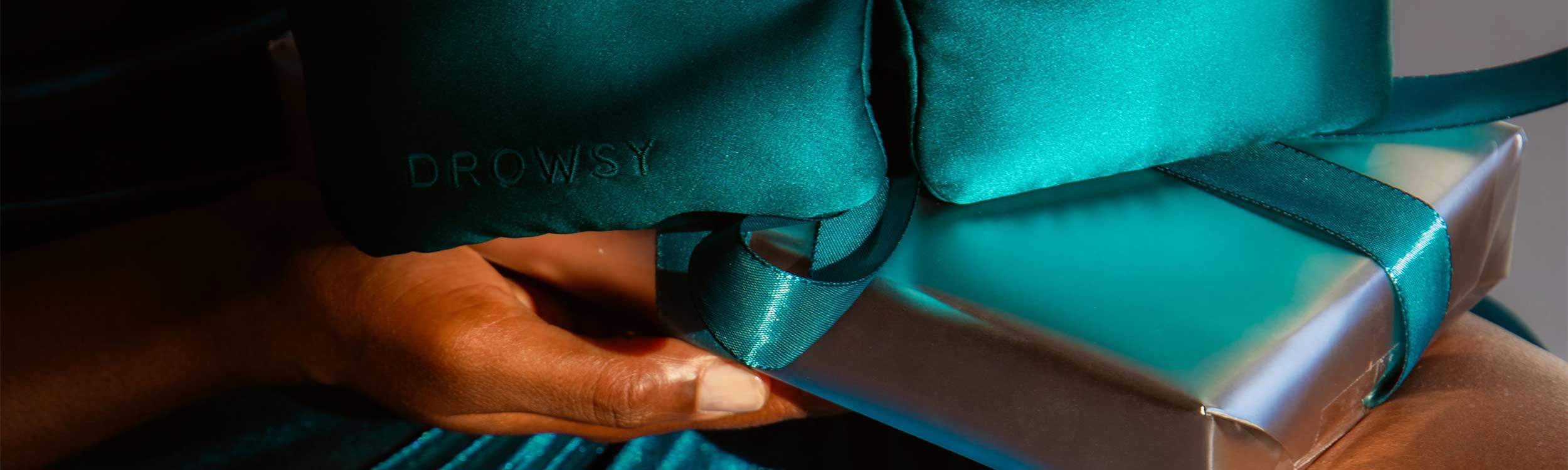 A hand holds a silver gift box wrapped with a teal ribbon. Resting on top is a teal satin eye mask with the word DROWSY embroidered on it. The scene is lit warmly, highlighting the shiny textures.