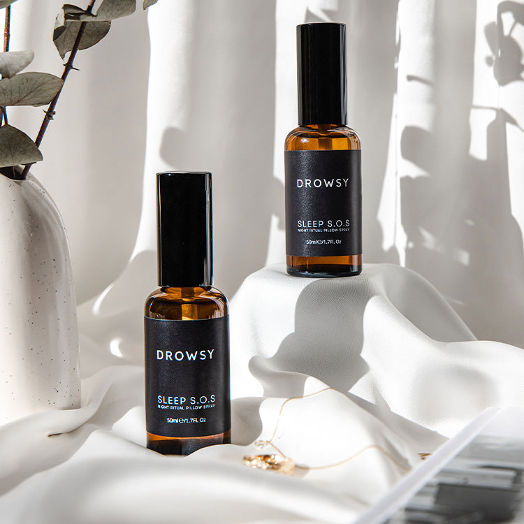 Two amber glass spray bottles labeled Drowsy Sleep S.O.S sit on a white, fabric-draped surface with dried eucalyptus branches and soft natural light casting shadows in the background.