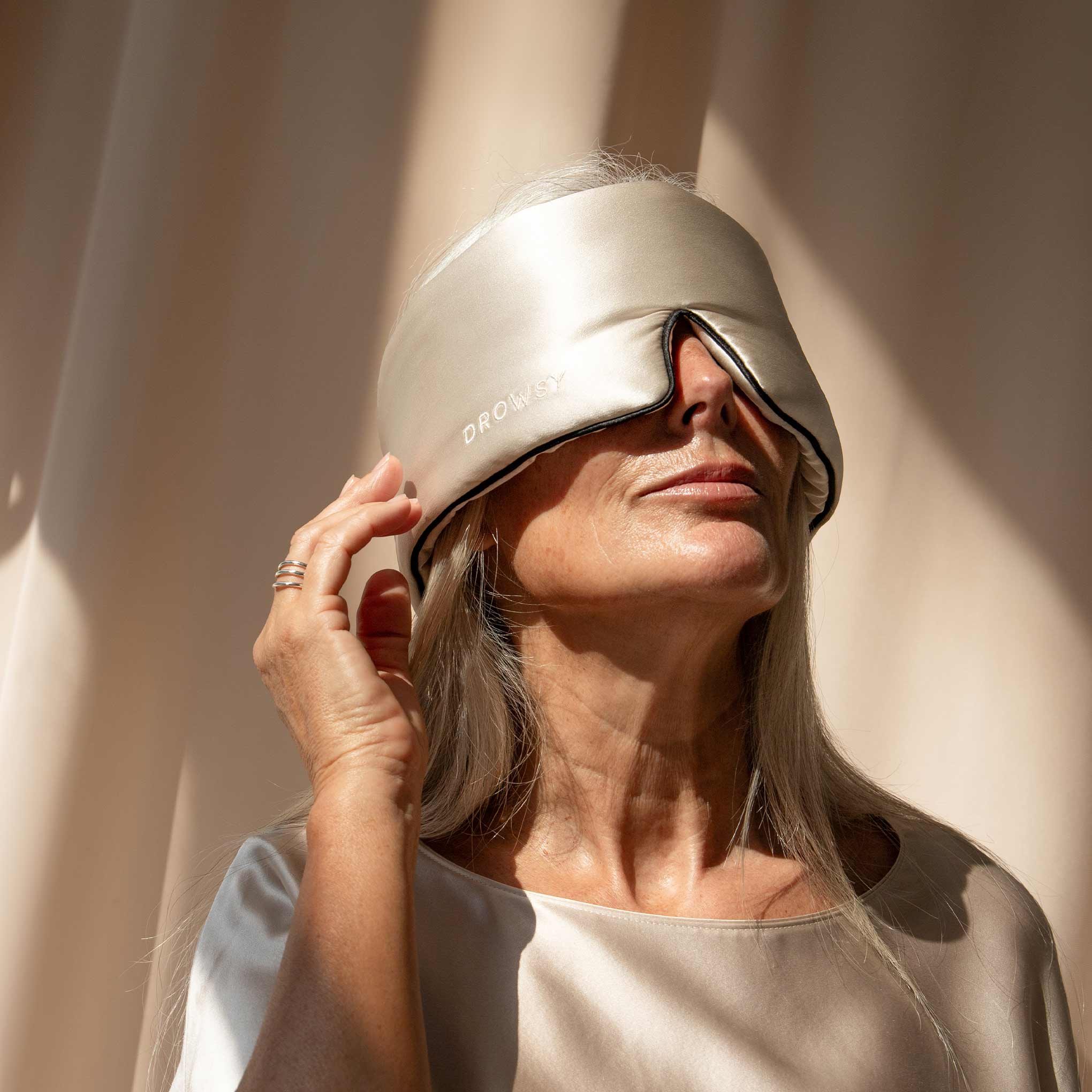 An older woman with long gray hair wears the Dusty Gold - Pipe Dreams Edition sleep mask by us.drowsysleepco, labeled DROWSY. She gently touches the edge in soft light. The washable mask features an adjustable strap for comfort.