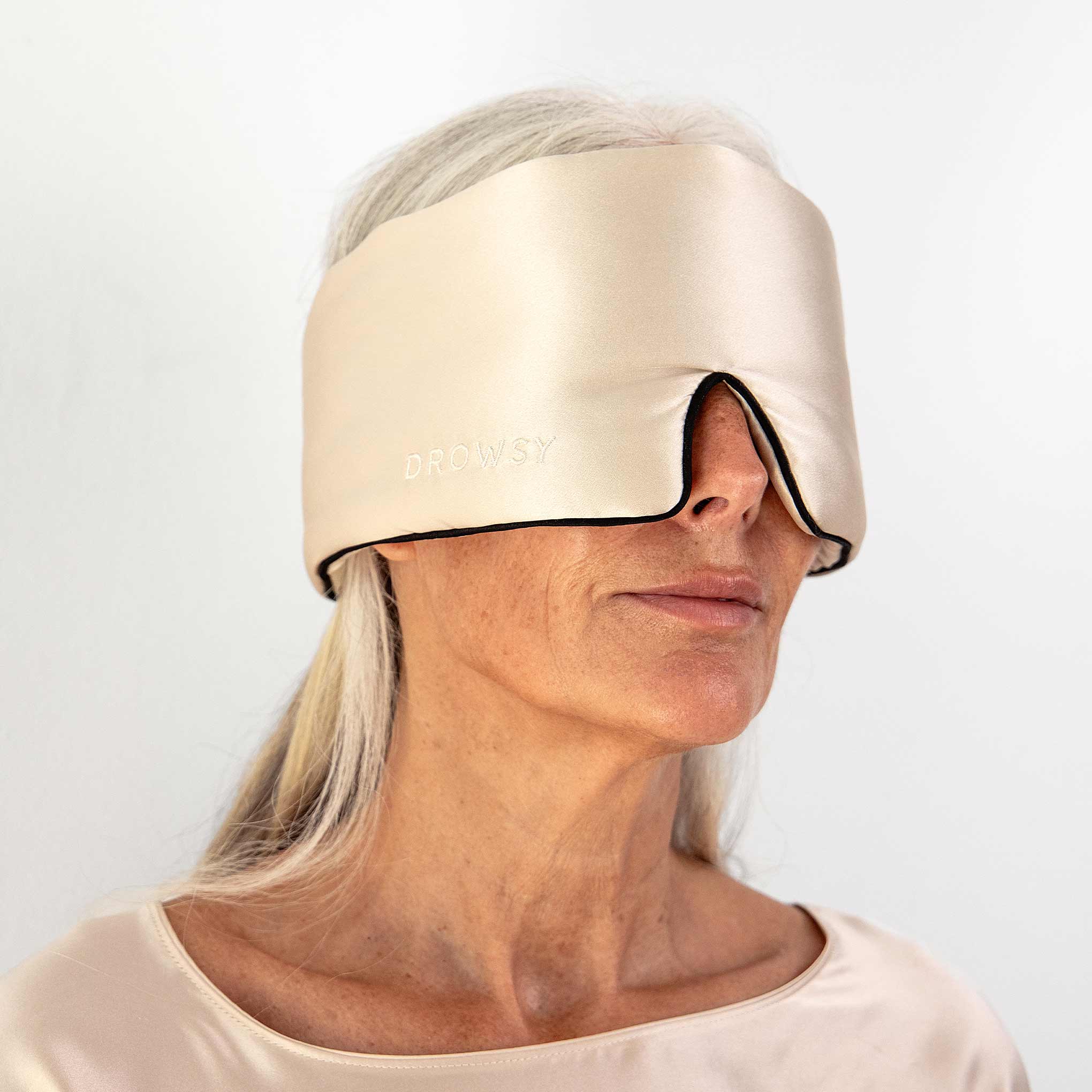 An older woman with long gray hair wears the Dusty Gold - Pipe Dreams Edition sleep eye mask by us.drowsysleepco, made of washable mulberry silk, covering her eyes and nose. She sports a matching satin top and adjustable head strap against a white background.