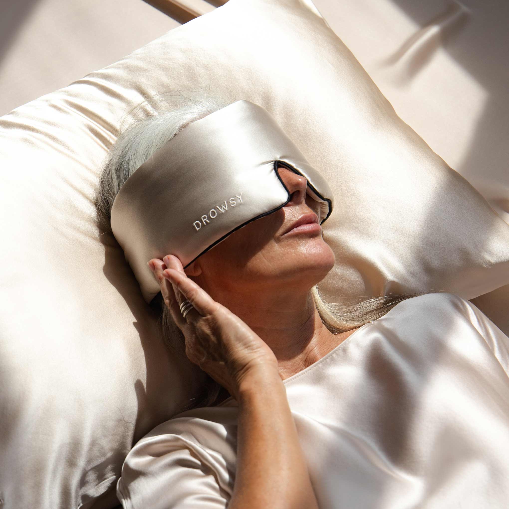 A woman with long gray hair lies on a satin pillow, wearing the Dusty Gold - Pipe Dreams Edition sleep mask by us.drowsysleepco. Dressed in a matching satin top, she rests her hand on the mask as sunlight filters onto the bed.