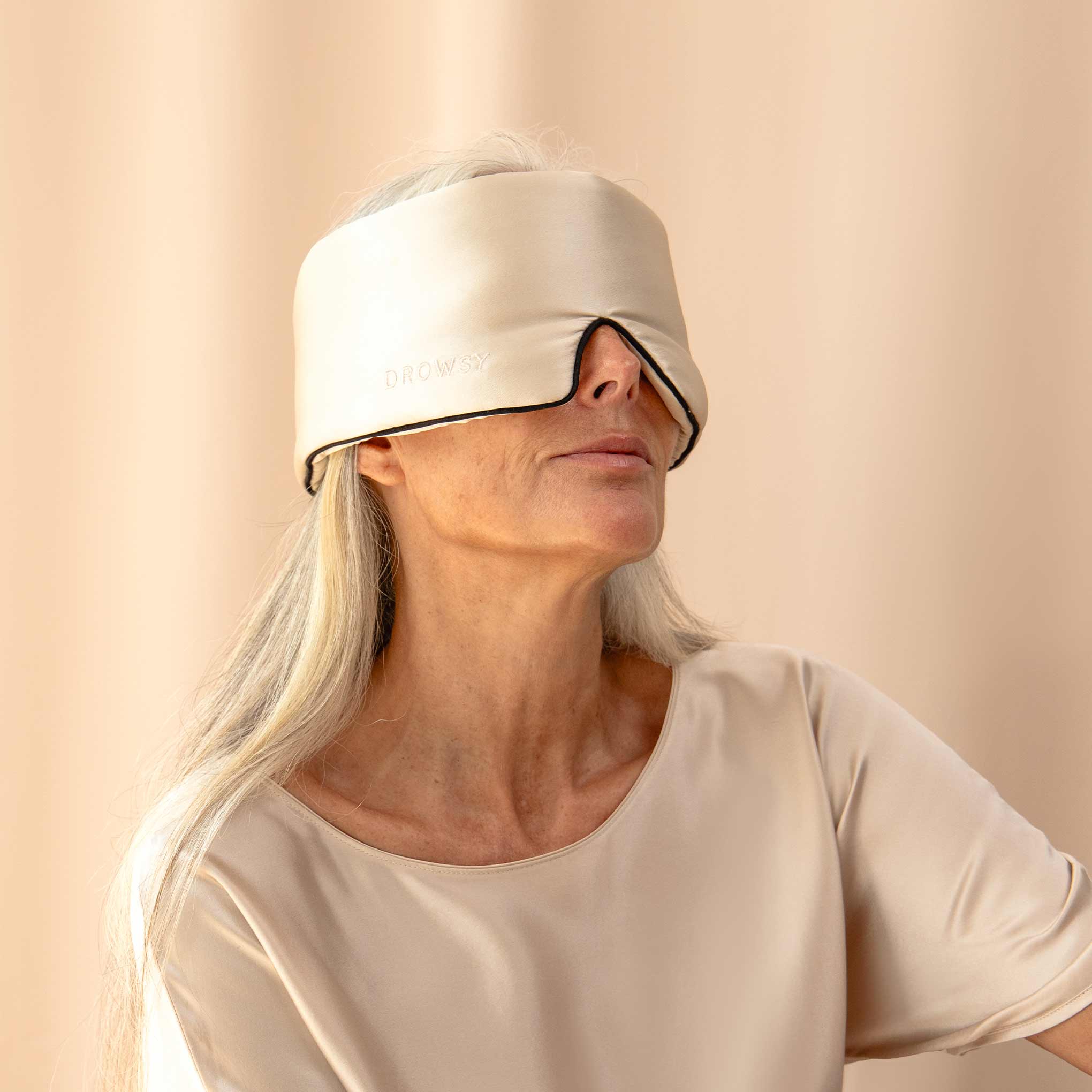 A woman with long gray hair wears the us.drowsysleepco Dusty Gold - Pipe Dreams Edition mulberry silk sleep mask with an adjustable strap, paired with a beige top, standing against a soft beige background.