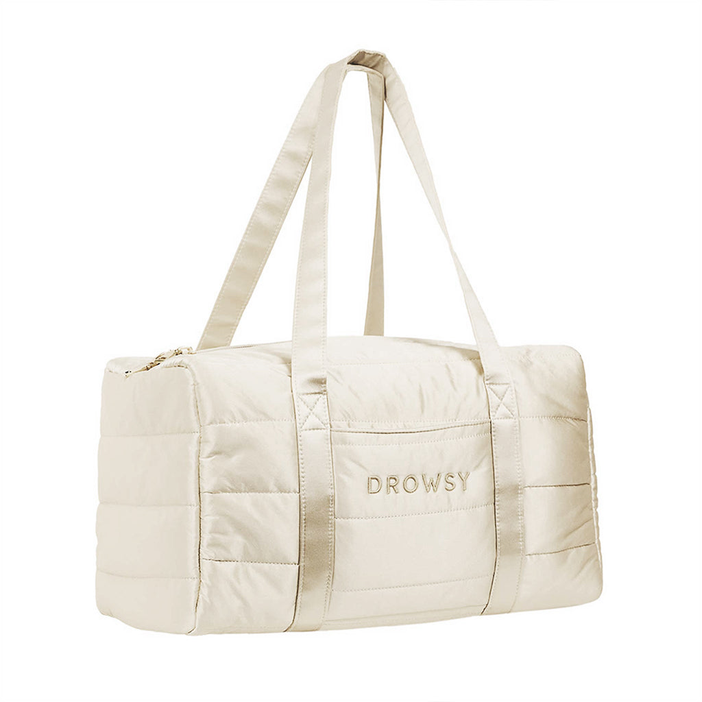 The Drowsy Silk Weekender in Dusty Gold by Drowsy is a cream-colored, quilted travel bag with two handles and "DROWSY" embroidered on the front pocket—perfect for your next weekend getaway.