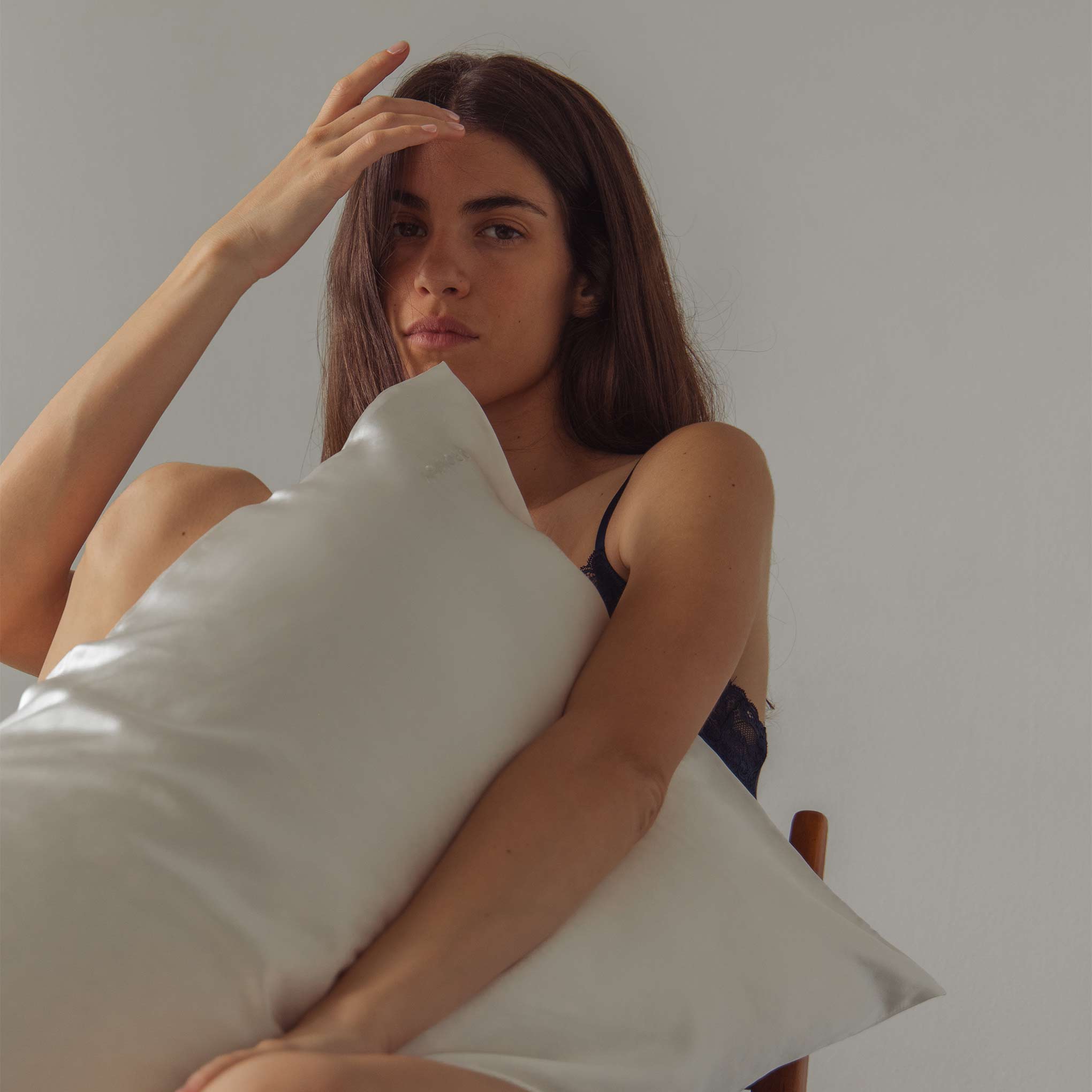 A woman with long brown hair sits on a chair, holding the drowsysleepco Perfect Skin Bundle Midnight Blue silk pillowcase. Wearing a dark sleeveless top, she gazes at the camera with one hand on her forehead against a plain light background.