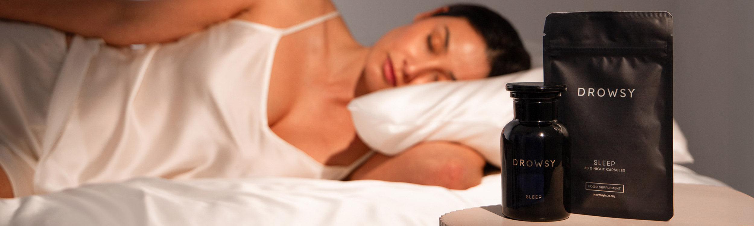 A woman in a white satin slip sleeps on a bed. In the foreground, two black Drowsy branded sleep supplement products are placed on a nightstand.