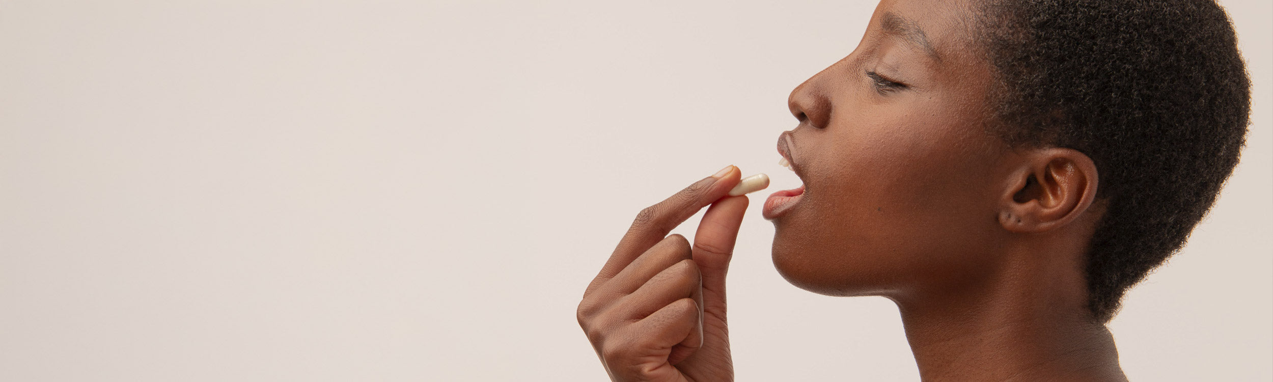 A person with short hair holds a white capsule near their mouth, preparing to take it, against a plain light background.