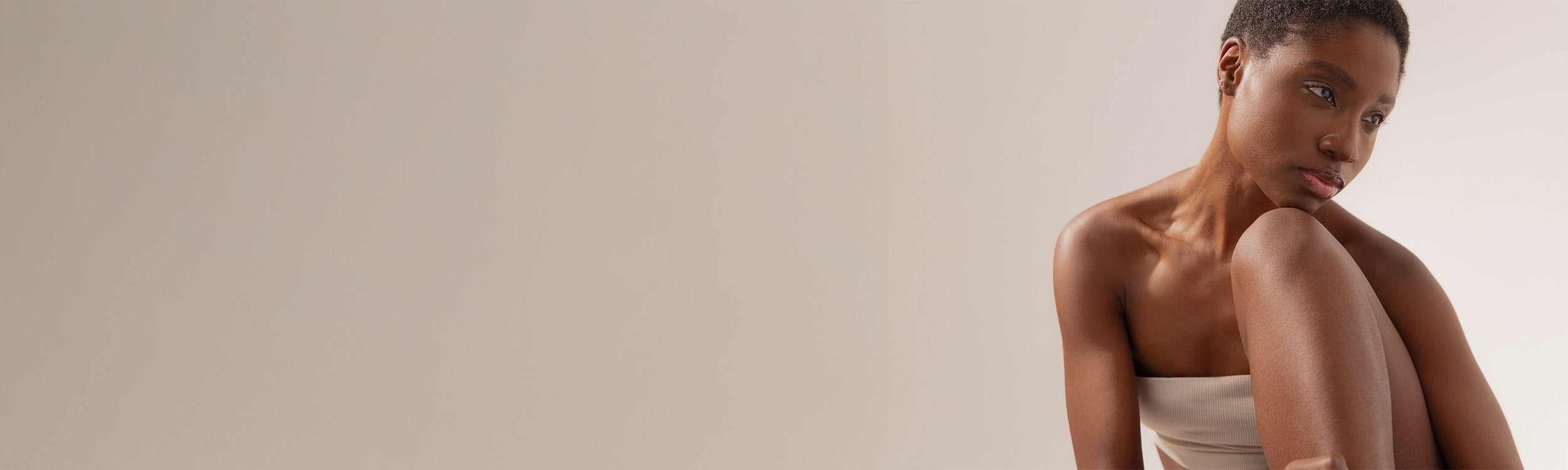 A woman with short hair sits with her knees drawn up, wearing a strapless beige top, against a plain, light background. She gazes thoughtfully downward, with soft lighting highlighting her features.